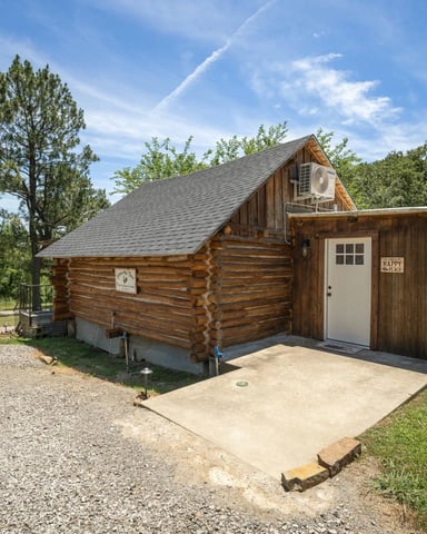 Small log cabin with a shingled roof and white door beside a gravel drive